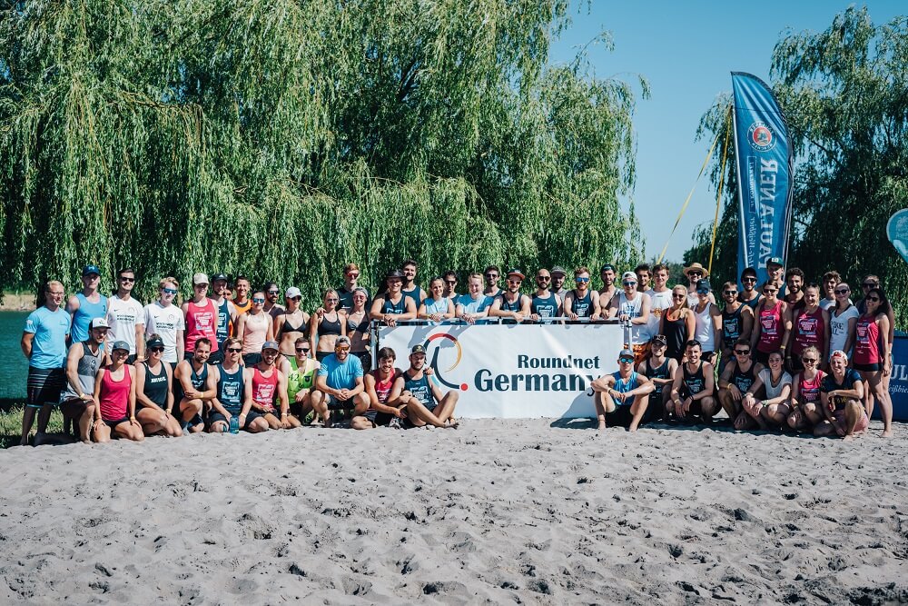 Ca. 50 Menschen posieren für ein Gruppenfoto auf Sand